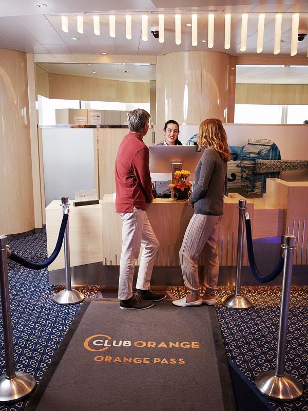 Guests receiving concierge services at the Club Orange priority desk on a Holland America Line cruise ship, standing on a branded 'CLUB ORANGE ORANGE PASS' mat, indicative of the exclusive experience offered.
