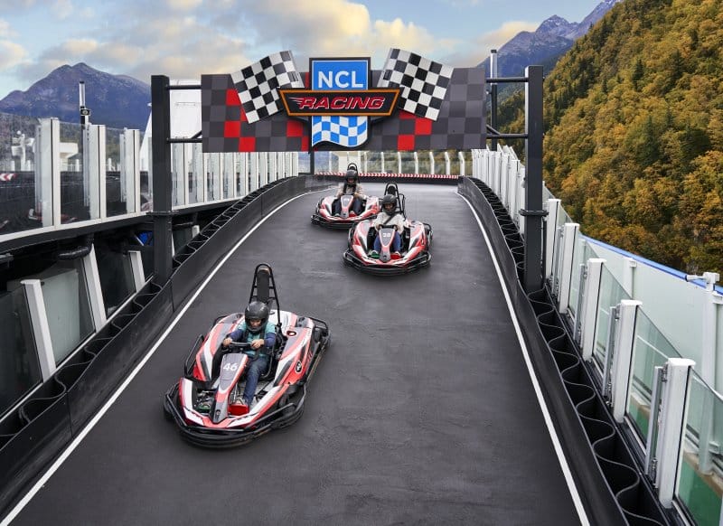 Passengers in go-karts race on the NCL Racing track atop the NCL Bliss cruise ship, with a stunning mountainous backdrop and lush greenery surrounding them, offering an exhilarating and scenic on-board activity.