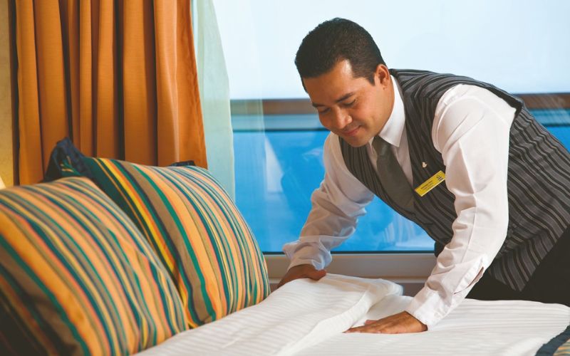A crew member of Costa Cruises attentively making a bed, with a focus on comfort and neatness, showcasing the detailed and personal care provided onboard, complemented by the warm interior colors and natural light from the cabin window.