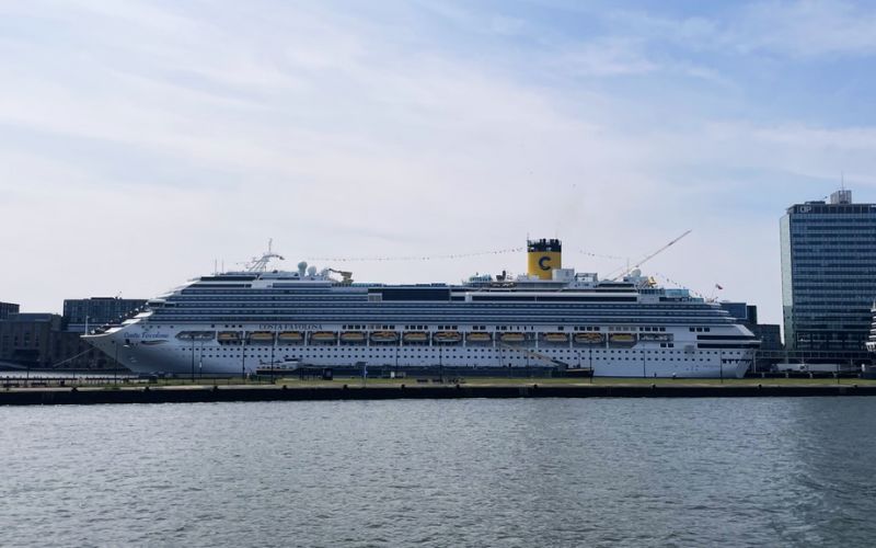 The Costa Favolosa cruise ship moored in Amsterdam, with its distinctive yellow funnel and multiple decks, set against the backdrop of the city's modern buildings and the calm waters in the foreground.