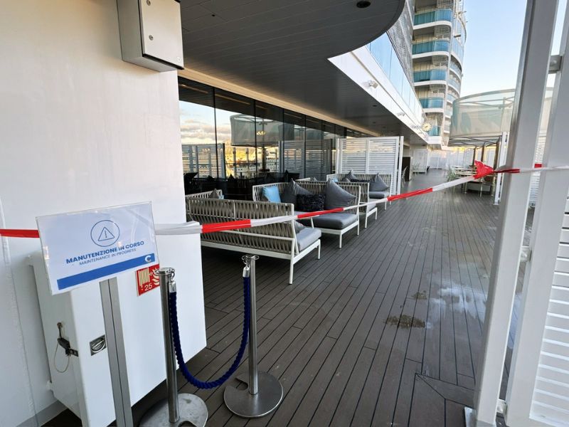 An area of the Costa Smeralda's deck is cordoned off for maintenance, with a sign reading 'Manutenzione in corso,' alongside comfortable outdoor lounge seating, with the early evening sky reflecting off the glass panes above.
