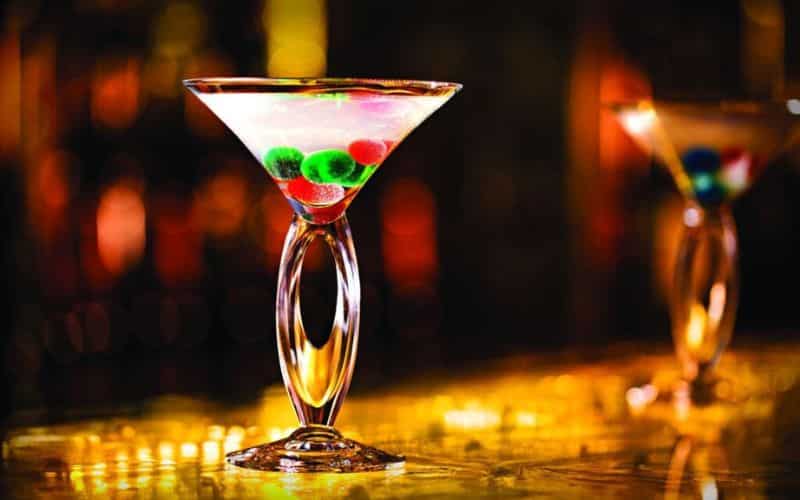 Two elegant martini glasses with a unique twisted stem design, the forefront glass filled with a clear liquid and colorful spherical garnishes, against a warm, bokeh-lit bar backdrop.