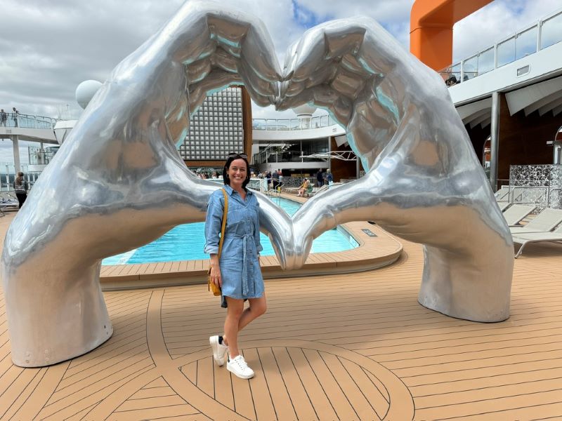 Cruise Mummy in a light blue dress stands in front of a large silver sculpture of two hands forming a heart shape on a Celebrity Cruises ship. The deck features a small pool in the background, and the woman smiles at the camera under a partly cloudy sky.