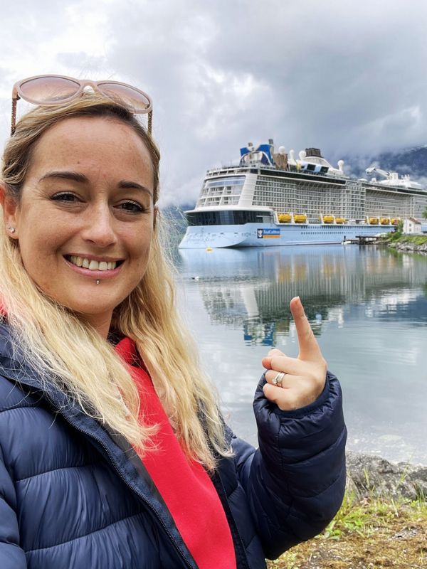 Cruise Mummy in a blue puffer jacket and pink hoodie points and smiles at a Royal Caribbean cruise ship docked in a scenic, misty fjord. The ship's reflection is visible in the calm water, with mountains and cloudy skies in the background.