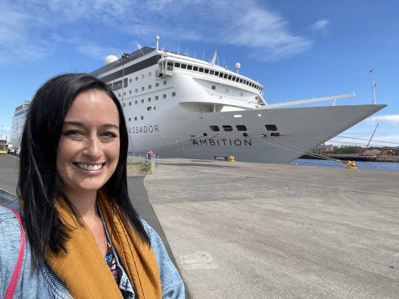 Cruise Mummy with long dark hair smiles at the camera, standing in front of the Ambassador Cruise Line's ship named Ambition docked at a port. The ship is large and white with the words "AMBASSADOR CRUISE LINE" and "AMBITION" clearly visible on its hull.