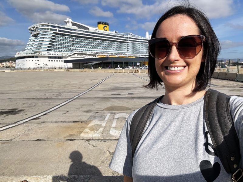 Cruise Mummy wearing sunglasses and a gray t-shirt with a black heart symbol stands smiling in front of a large Costa Cruises ship docked at a port. She has a backpack on, and the ship is visible against a partly cloudy sky.