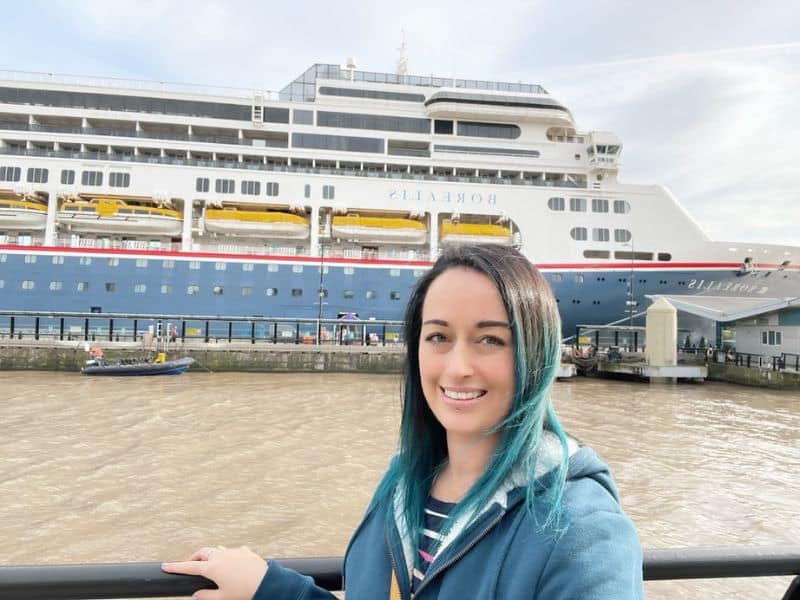 Cruise Mummy with teal highlights in her hair smiles while standing in front of the Fred. Olsen Borealis cruise ship docked at a port. The ship's name "Borealis" is visible on the side, and the woman is wearing a blue jacket with a striped shirt underneath.