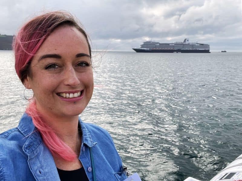 Cruise Mummy with pink highlights in her hair smiles while standing near the water with a Holland America Line cruise ship visible in the distance. She is wearing a denim jacket over a black top, and the cloudy sky adds a dramatic backdrop to the scene.