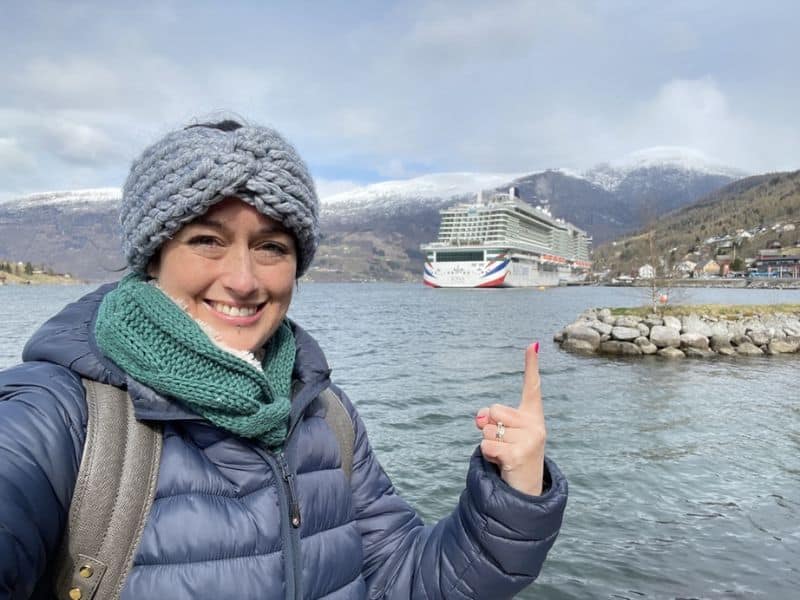 Cruise Mummy bundled up in a gray knit headband, green scarf, and blue puffer jacket smiles while pointing at a P&O cruise ship docked in a scenic fjord. Snow-capped mountains and quaint buildings create a picturesque background under a partly cloudy sky.