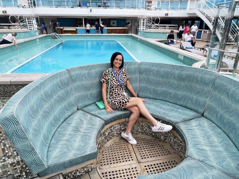 Cruise Mummy in a leopard print dress sits on a curved, patterned bench near a pool on a Princess cruise ship. She smiles at the camera, with people in the background enjoying the pool area under a covered deck.