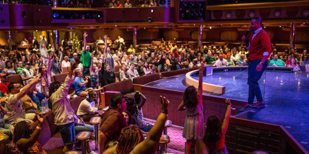 A Carnival cruise director is engaging with a lively audience in a large theater on the ship. The director, holding a microphone, stands on stage as numerous audience members raise their hands enthusiastically. The theater is filled with passengers seated in comfortable chairs, enjoying the interactive entertainment under warm, vibrant lighting.
