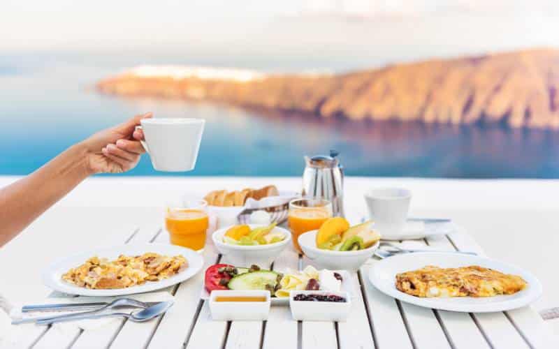 A  breakfast spread on a white table with a scenic ocean view on a cruise ship, featuring a hand holding a white coffee cup, plates of scrambled eggs and omelet, bowls of fresh fruit and yogurt, with orange juice and coffee on the side.
