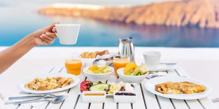 A breakfast spread on a white table with a scenic ocean view on a cruise ship, featuring a hand holding a white coffee cup, plates of scrambled eggs and omelet, bowls of fresh fruit and yogurt, with orange juice and coffee on the side.