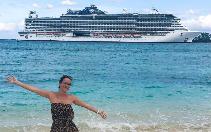 A smiling woman in a leopard print dress with sunglasses on her head, extends her arms joyfully on a sandy beach with the MSC cruise ship in the background, floating on calm turquoise waters under a partly cloudy sky.
