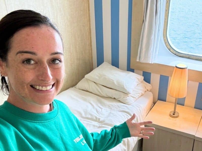 This image features a smiling Cruise Mummy in a green sweatshirt posing in a cosy cabin with two twin beds separated by a small nightstand. The nightstand holds two warm-lit lamps, and a large window in the background offers a view of the ocean. The striped blue and white wallpaper adds a nautical touch to the room.