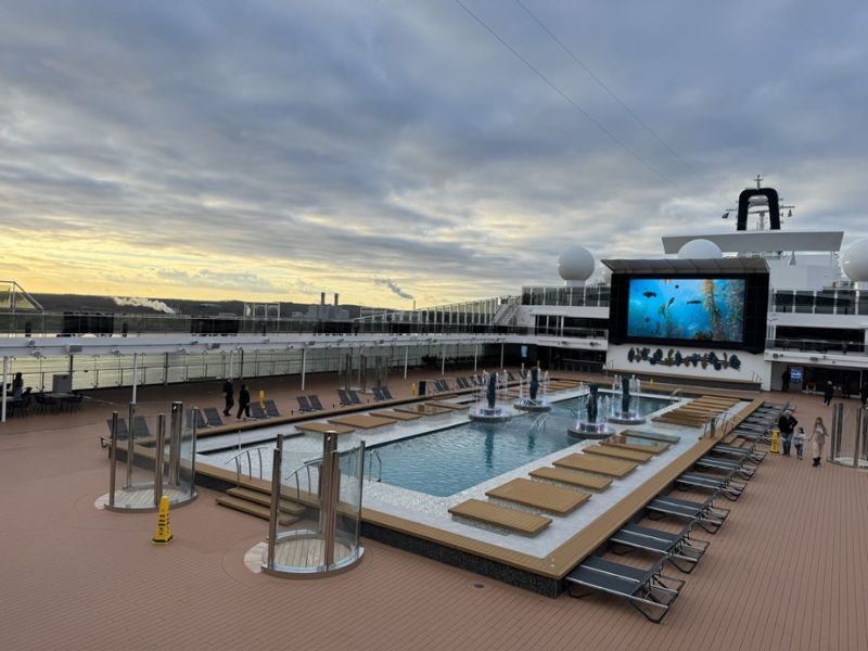 Early evening on a cruise ship's pool deck, featuring loungers, a tranquil pool with fountains, and a large outdoor movie screen displaying underwater imagery. The deck overlooks a port with industrial structures in the distance, under a soft sunset sky.