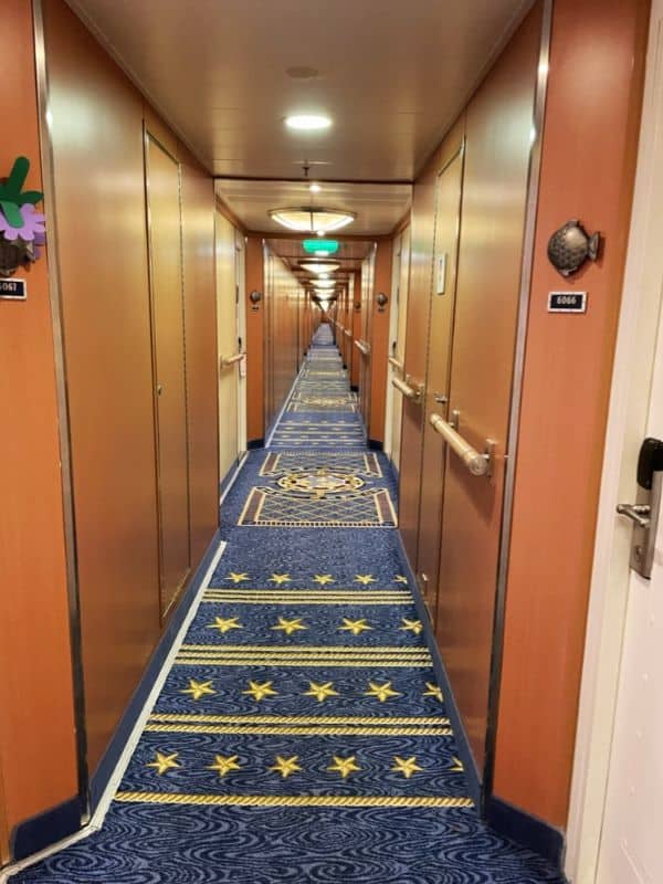 A long, narrow corridor onboard a cruise ship with patterned blue carpet featuring star motifs, flanked by cabin doors with decorative fish plaques, suggesting a theme that evokes the oceanic environment of the journey.