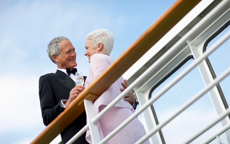 An older couple shares a tender moment on a cruise ship deck, elegantly dressed with the man in a tuxedo and the woman in a pink jacket, both smiling and holding champagne glasses, epitomizing timeless romance and luxury travel.