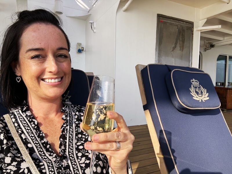 Cruise Mummy smiles at the camera while holding a glass of champagne on the deck of the Cunard Queen Mary cruise ship. She is sitting on a blue lounge chair with a regal emblem embroidered on the headrest.