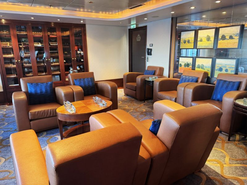 A cozy lounge area on the Cunard Queen Mary cruise ship features several plush leather armchairs arranged around small tables with glass ashtrays. The space includes a wall of wooden cabinets displaying a collection of cigars and other items, creating an inviting atmosphere for relaxation.