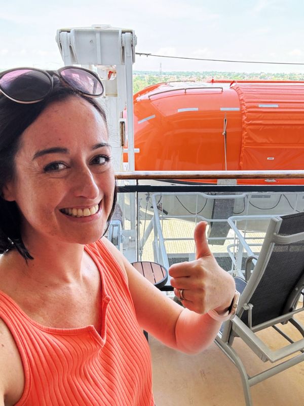 Cruise Mummy in an orange top gives a thumbs-up while sitting on the deck of the Cunard Queen Mary cruise ship. Behind her, an orange lifeboat is visible, along with some outdoor seating and railings. She appears to be enjoying her time on the ship.