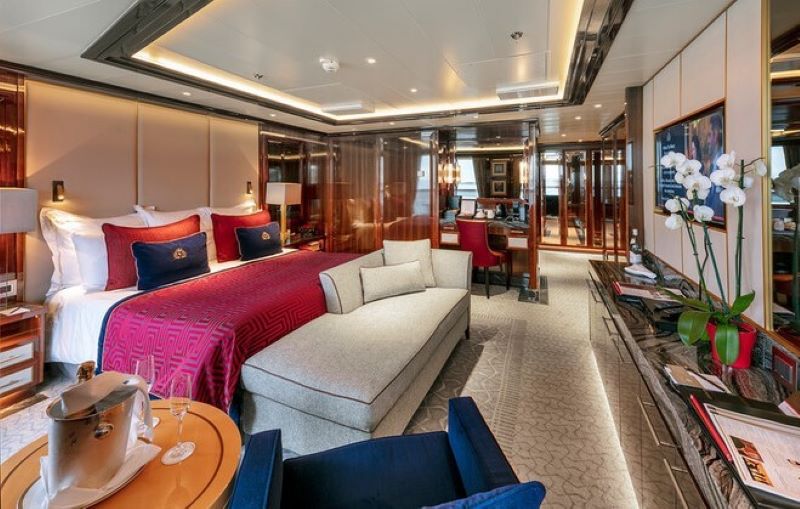Interior of a Cunard Cruise Queens Grill suite showcasing opulent decor. A king-sized bed adorned with a geometric-patterned red throw and navy blue pillows is centered in the room, complemented by a plush beige sofa and a glass-topped table with a champagne ice bucket and glasses. Rich wood paneling and an orchid arrangement add to the room's luxurious ambiance.