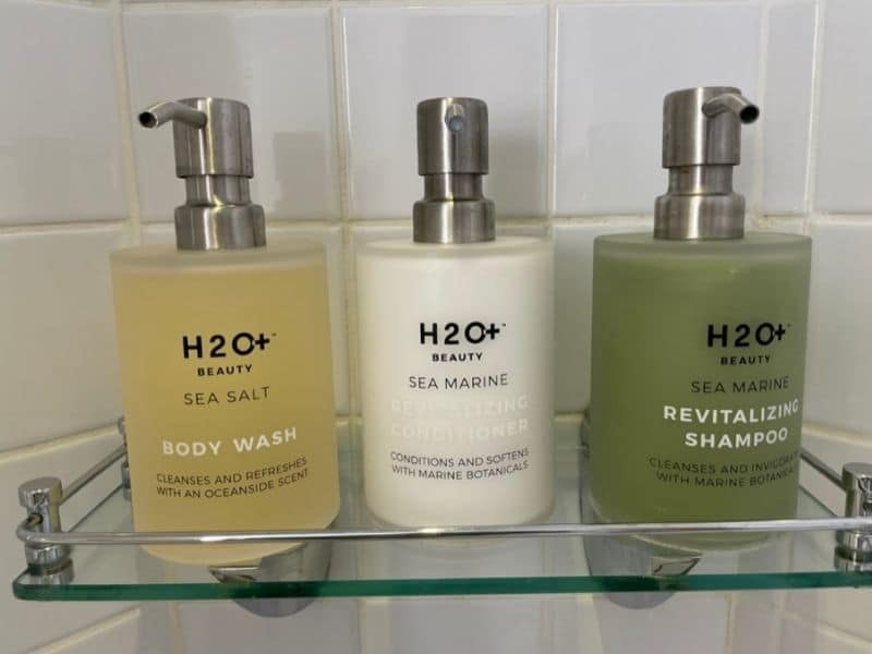 This image displays three H2O+ Beauty products arranged on a glass shelf in a tiled bathroom on a Disney cruise ship. From left to right, the products include a Sea Salt Body Wash, a Sea Marine Revitalizing Conditioner, and a Sea Marine Revitalizing Shampoo. Each bottle has a pump dispenser and is labeled with text highlighting marine botanicals and ocean-inspired scents.