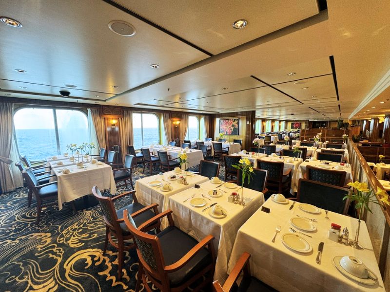 A spacious dining room on a Cunard cruise ship, featuring elegantly set tables with white tablecloths, flower centerpieces, and large windows offering an ocean view.