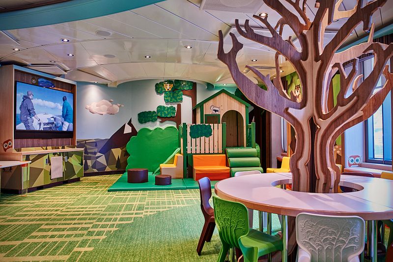 Camp Discovery children's play area on a Princess Cruise ship, showcasing a colorful, nature-themed indoor playground with a large tree structure, playful furniture, and a video screen for entertainment.