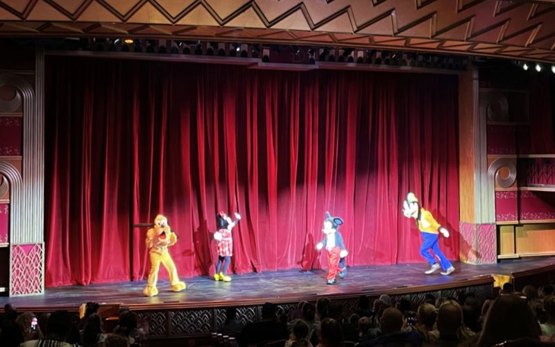 Disney Cruise Line entertainment in full swing with iconic characters Pluto, Minnie Mouse, Mickey Mouse, and Goofy delighting the audience during a lively performance on stage.