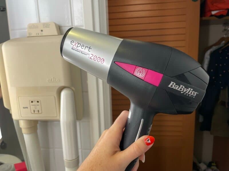 A hand holds a BaByliss Paris hair dryer labeled "expert 12000" inside a cabin on the Disney Magic cruise ship. The background shows a wall-mounted hair dryer unit and a partially open closet with clothing.