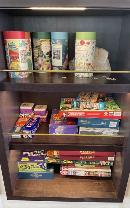 A wooden shelf stocked with a variety of classic board games such as Monopoly, Scrabble, Clue, and Charades, alongside decorative cylindrical tins, suggesting a cozy entertainment area on a cruise ship or in a vacation home.