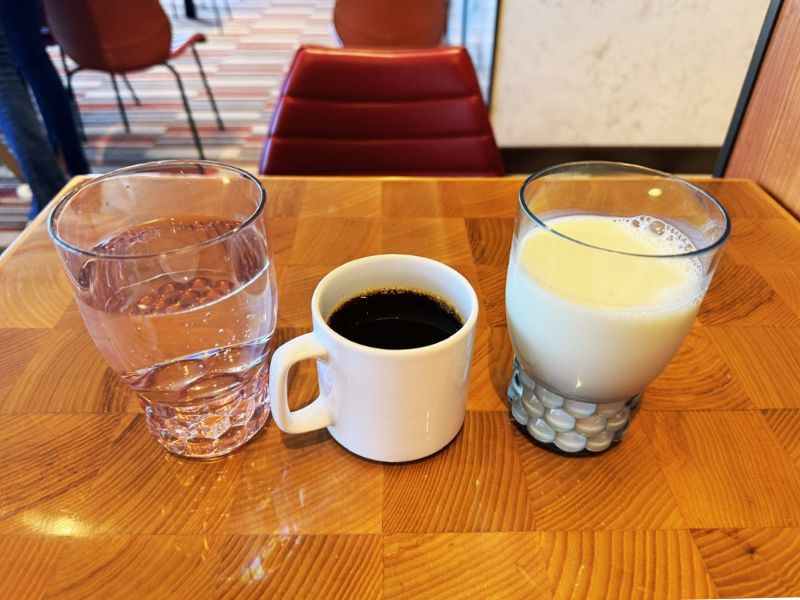 On a polished wooden table aboard the Costa Smeralda, a refreshing set of drinks is presented: a glass of water, a mug of black coffee, and a glass of milk, against the backdrop of the ship's cozy interior seating area.