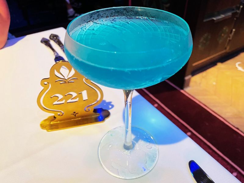 A striking bright blue cocktail served in a classic stemmed glass, showcased on a table aboard a Disney cruise ship. The vibrant blue color of the drink adds a playful and exotic touch, making it a visual standout. Accompanying the cocktail is a golden placard displaying the number "221", likely denoting a table or room number. The table setting, accentuated by the deep hues of the drink and the elegant gold placard, creates a luxurious and inviting ambiance typical of Disney cruise line's attention to detail in their dining experiences.