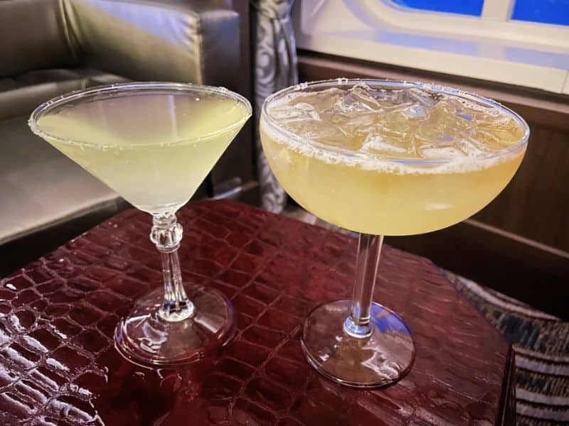 Two cocktails on a Princess cruise ship placed on a red textured table. One drink is in a martini glass with a light green color, and the other is in a coupe glass filled with ice and a light yellow beverage. A window and part of a seating area are visible in the background.