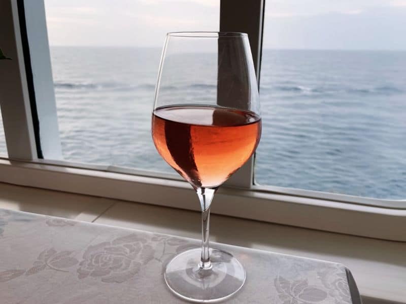 A glass of rosé wine sits on a table near a window on a Princess cruise ship, with the ocean visible in the background. The scene captures a calm, scenic view of the sea, enhancing the relaxing ambiance.