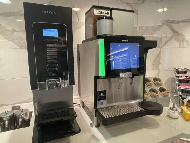 A drink station on a Princess cruise ship featuring two machines: one labeled "OptiVend" and the other for regular coffee with a touch screen interface. The area is clean and modern, with condiments and cups neatly arranged beside the machines.