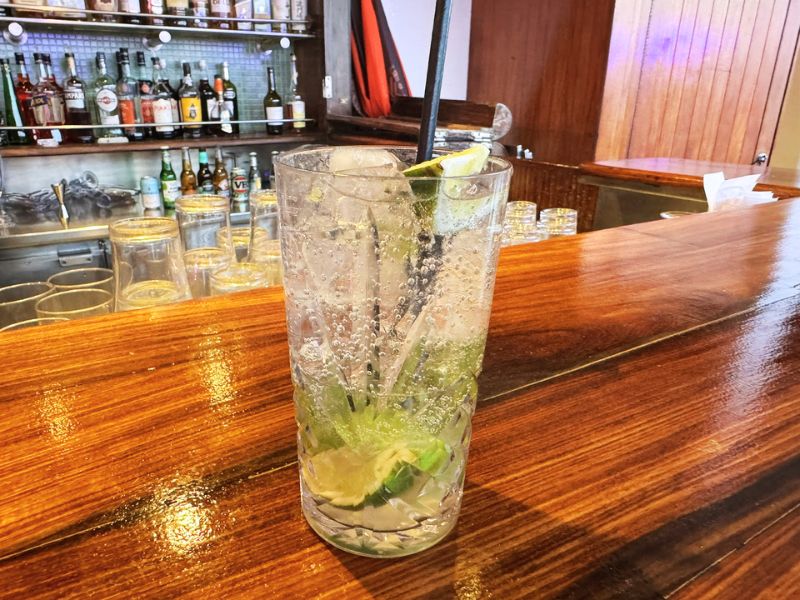 A Mojito cocktail with lime and ice sits on a wooden bar counter on a Cunard cruise ship, with a background of various liquor bottles and glassware.