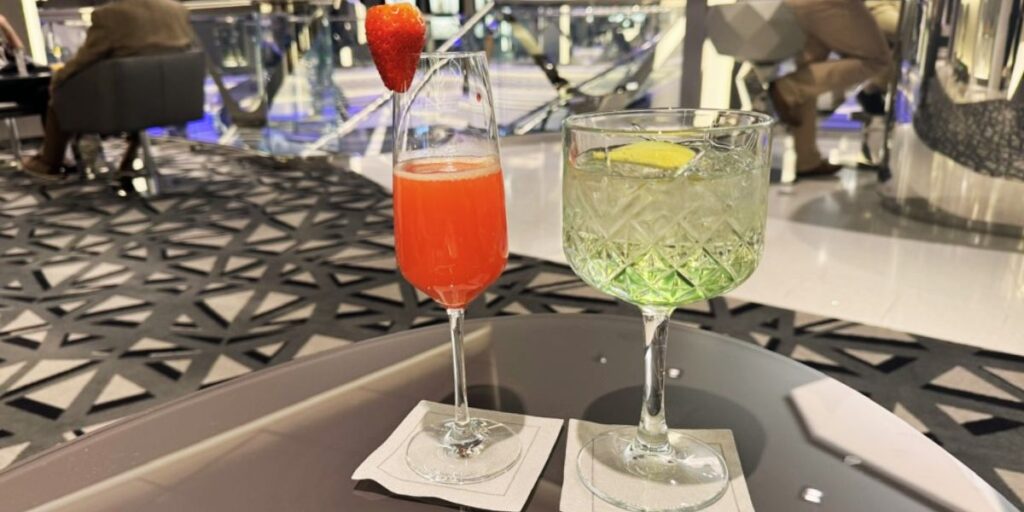 Two cocktails on a cruise ship table, one red with a strawberry garnish and the other green with a lemon slice. The setting features a modern, geometric-patterned carpet and stylish furniture, with passengers in the background enjoying the ambiance.