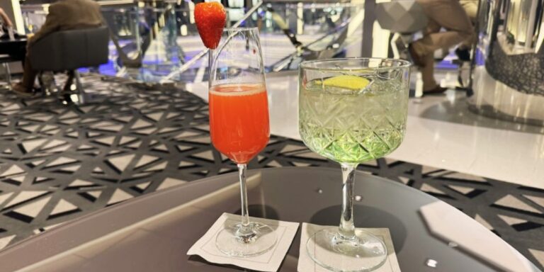 Two cocktails on a cruise ship table, one red with a strawberry garnish and the other green with a lemon slice. The setting features a modern, geometric-patterned carpet and stylish furniture, with passengers in the background enjoying the ambiance.