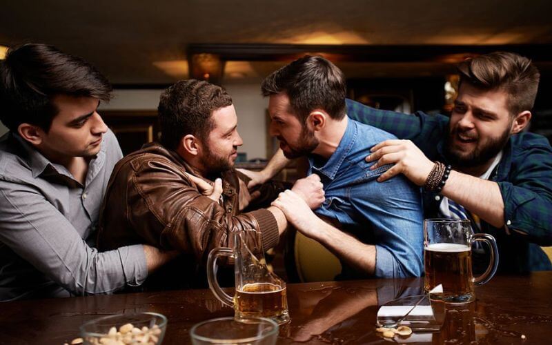 This image shows four men in a heated confrontation at a bar on a cruise ship. Two of the men in the center are grappling each other's shirts aggressively, while the other two on the sides attempt to pull them apart. There are beer mugs on the table in front of them, indicating they might be intoxicated.