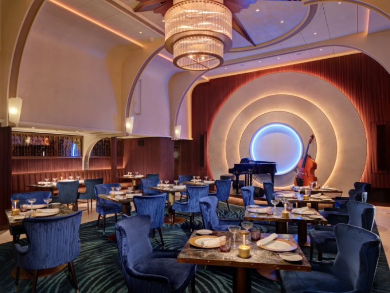 The Empire Supper Club on Royal Caribbean's Icon of the Seas exudes sophistication with its art deco-inspired décor, plush blue velvet chairs, and an illuminated circular stage with a grand piano, setting the stage for an elegant dining and live music experience.