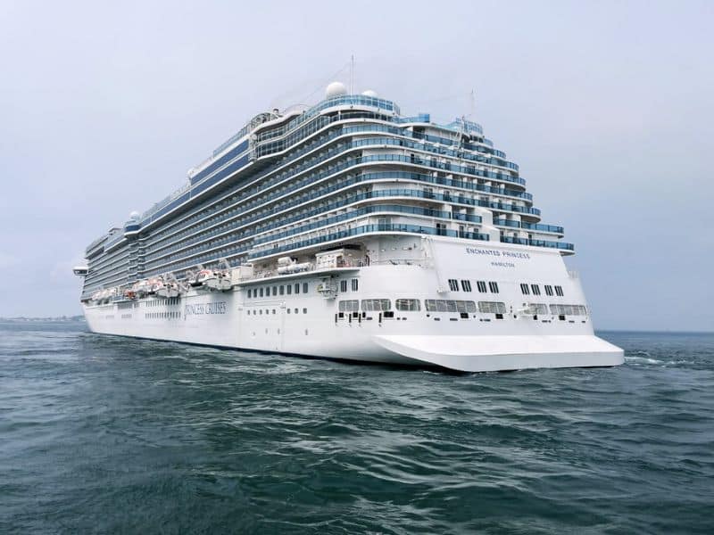 The Enchanted Princess cruise ship sailing on the ocean, showcasing its large white exterior with multiple decks and balconies. The ship's name and homeport, "Enchanted Princess" and "Hamilton," are visible on the stern. The sky is overcast, adding a soft light to the scene.