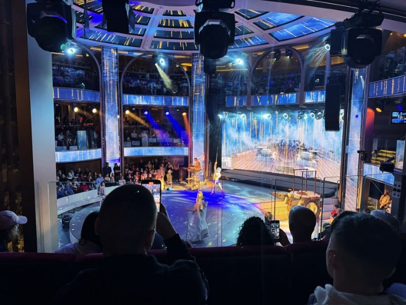 Passengers on the Costa cruise ship enjoy a vibrant live performance in the multi-tiered theater, with stage lights casting a glow on the performers and the audience immersed in the entertainment.