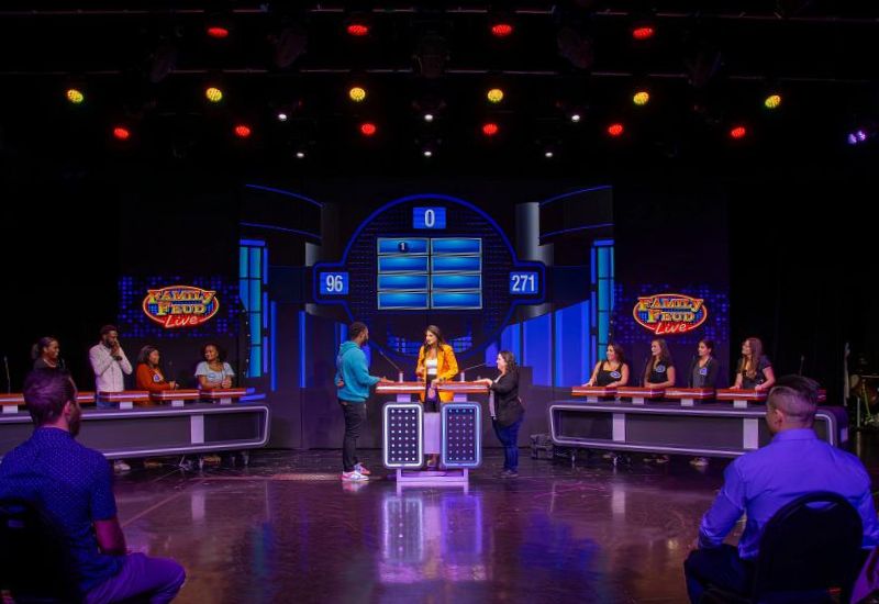 Two families face off in a lively game of 'Family Feud Live' on a Carnival cruise ship, set against a professional stage backdrop with colorful lights and scoreboards displaying their points. The audience, immersed in the excitement, watches as contestants eagerly anticipate the next question.