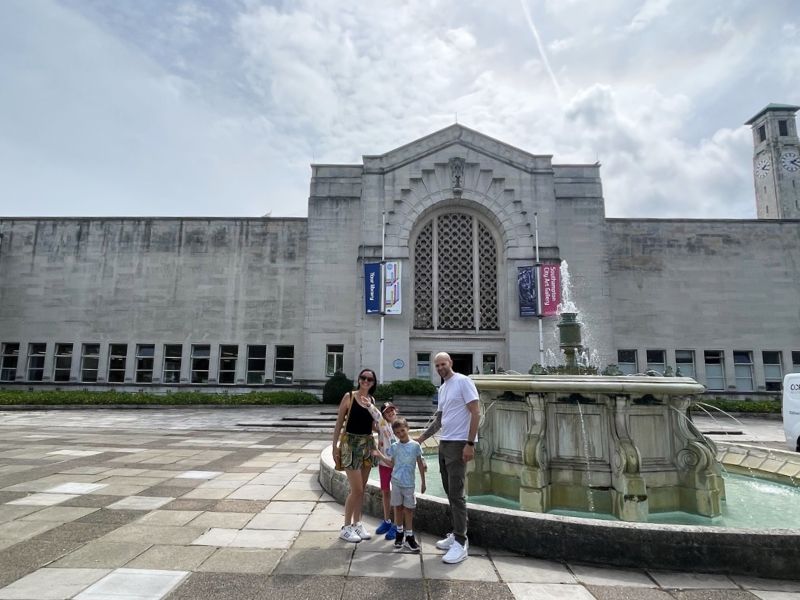 This image shows my family standing together near a fountain in front of Southampton City Art Gallery. The building's prominent facade features large arched windows and banners hanging from either side. The family appears to be enjoying a day out, with the child holding hands with both adults, creating a warm, relaxed atmosphere. The scene is bathed in soft daylight, with a partially cloudy sky overhead.