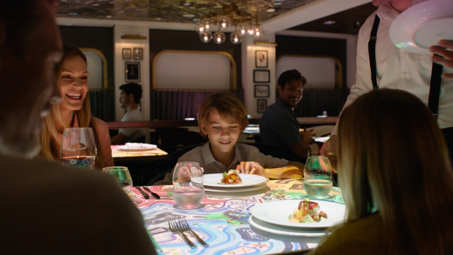 A family enjoys a meal together in a chic and modern dining area aboard a Celebrity cruise ship. The table is interactive with a vibrant, colorful pattern, and the young boy looks delighted with his dish, as the attentive server presents plates to the guests, creating a warm and memorable dining experience.