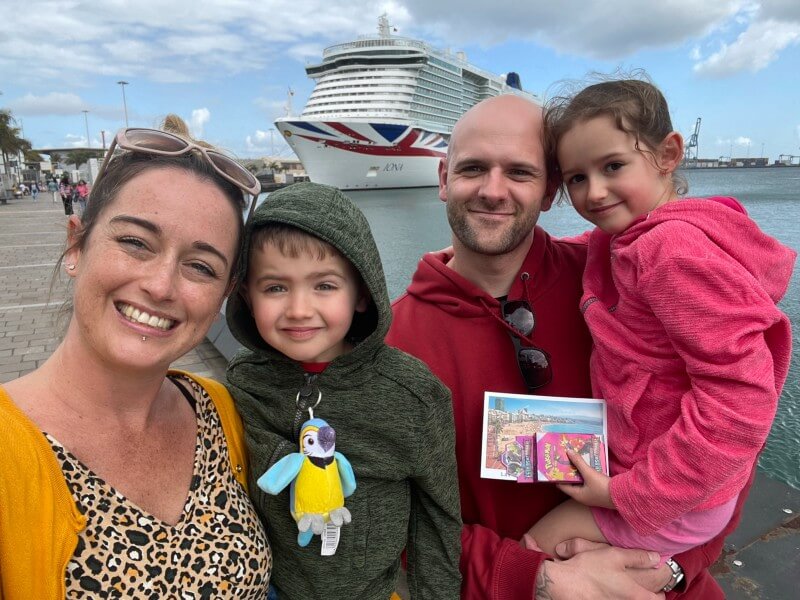 A smiling family of four stands in front of a docked P&O cruise ship. The mother, wearing a yellow cardigan, holds the camera, while the father, in a red hoodie, stands beside their son in a green hoodie and daughter in a pink jacket, who holds postcards. The cruise ship features the Union Jack on its bow.