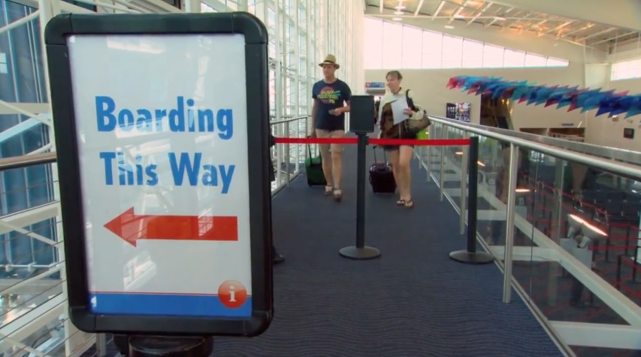 Passengers walking towards the priority check-in for a Carnival Cruise, guided by a sign that says 'Boarding This Way' with a red arrow, indicating the direction in a bright and airy terminal with glass walls and festive decorations.