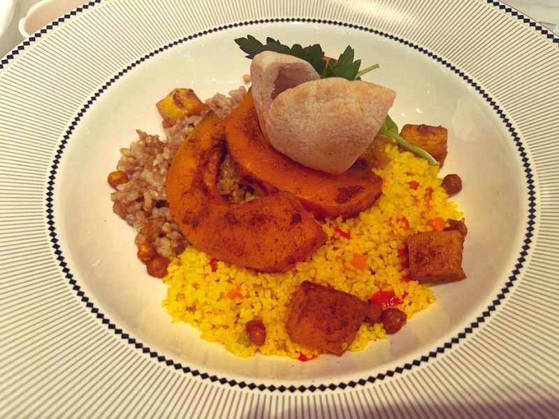 A beautifully plated meal served on a Disney cruise ship. The dish features a mound of vibrant yellow couscous surrounded by hearty chunks of spiced roasted pumpkin. Accompanying the couscous and pumpkin are garnishes of green herbs and several pieces of tofu, adding a protein component to the meal. A piece of bread, shaped like a small round bun, crowns the arrangement, offering a complete and visually appealing vegetarian option. The dish is served on a white plate with a classic blue striped rim, enhancing its visual appeal.
